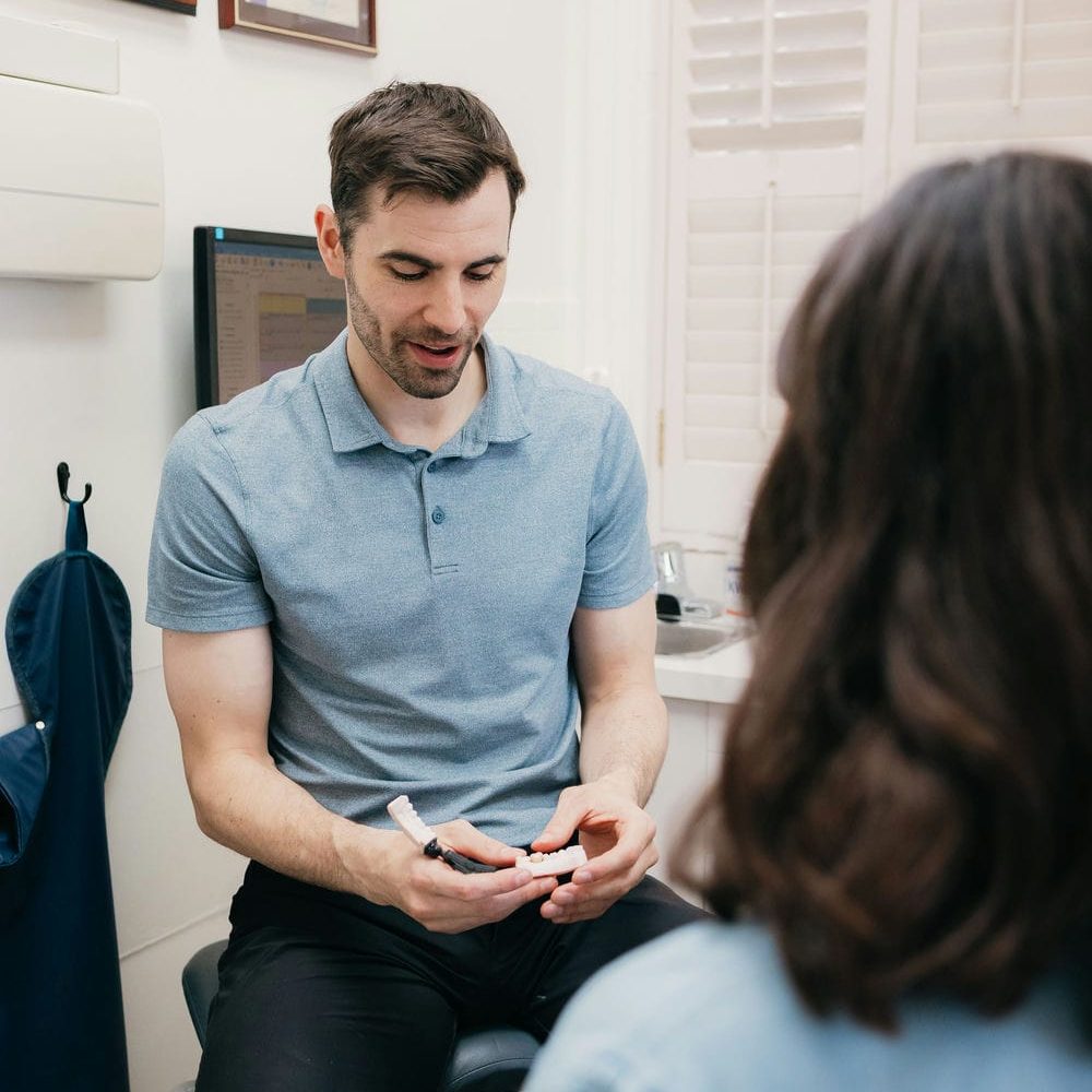 Dentist using a tooth model to explain the alternatives to a dental bridge to a seated patient