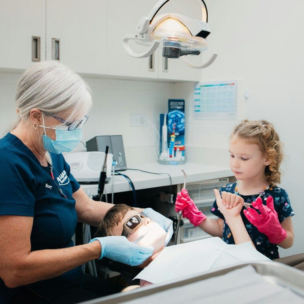 Hygienist treating a young boy while his little sister watches from beside the chair in oversized gloves