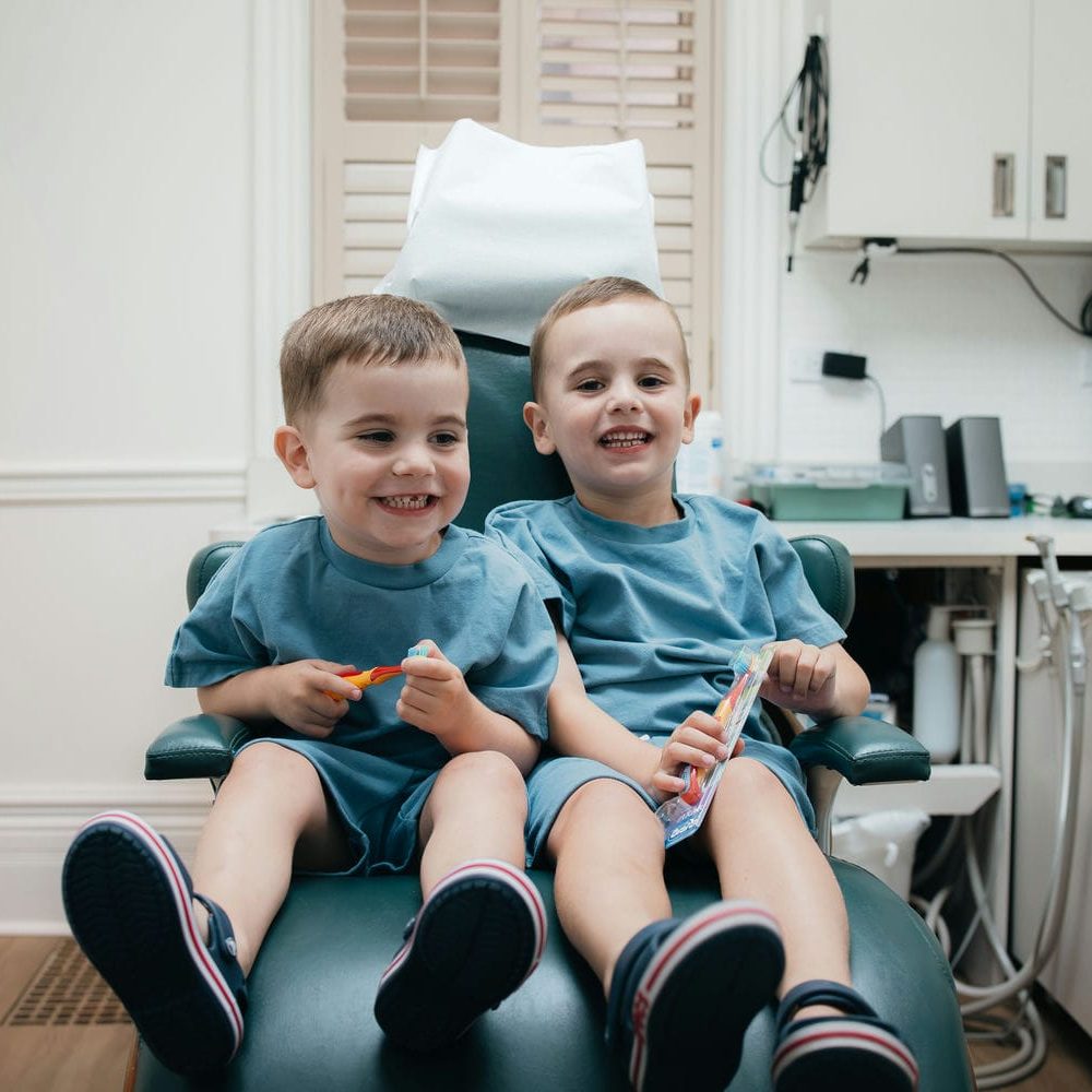 Two young brothers sitting together in the dental chair holding new toothbrushes after their checkups